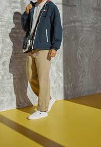 Marinblå huvmundjacka med vita dragkedjor, beige löst sittande byxor och vita sneakers. Lättviktigt tyg, avslappnad design, minimal detaljer.