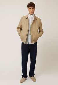 Chaqueta bomber beige claro de tejido texturizado, combinada con una camisa blanca y jeans azul oscuro, complementada con mocasines de color claro.