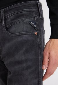 Sötétszürke farmernadrág slim fit szabású, enyhén kopott hatású, és díszes zsebcímke található rajta "REPLAY" felirattal. Réz szegecsek láthatók.