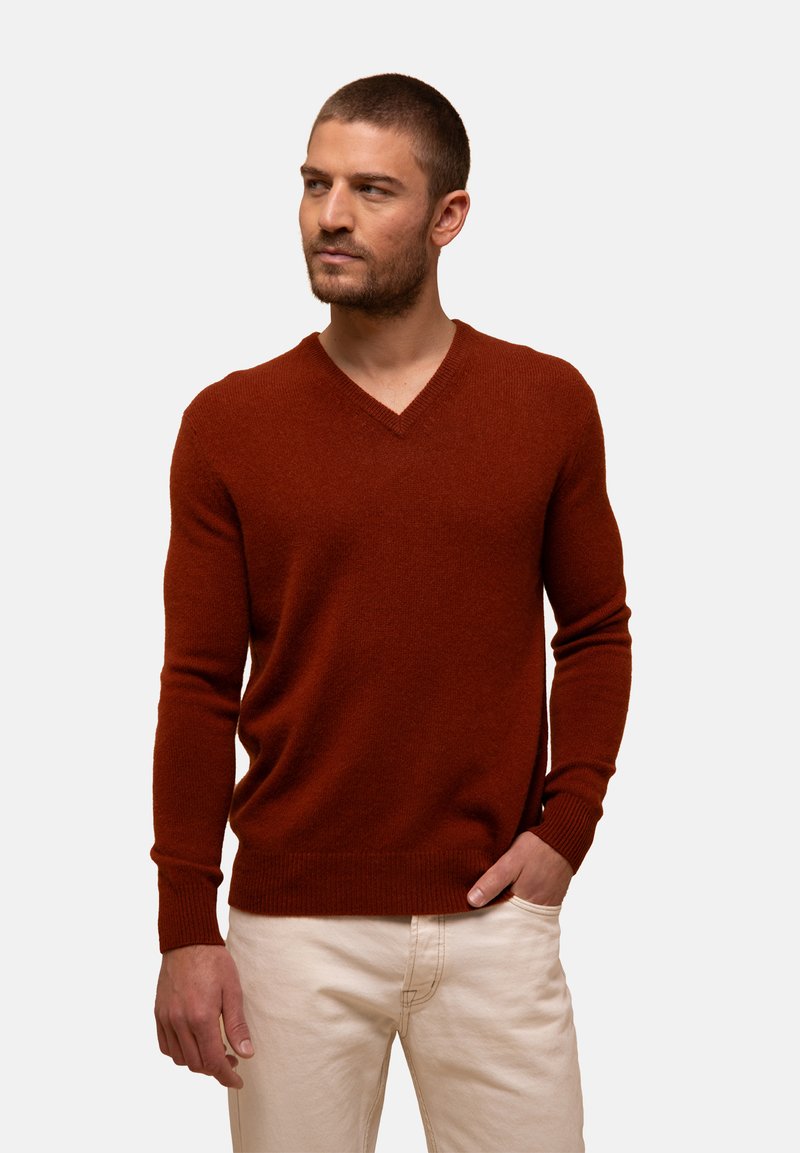 Rostfarbener V-Ausschnitt-Pullover aus weichem Material mit gerippten Bündchen und Saum, taillierter Schnitt, getragen über hellen Hosen.