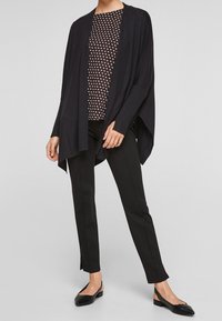 Black knit cardigan with open front, paired with a patterned blouse, black trousers, and black flats. Features loose, flowing design.