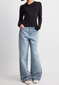 Top nero a maniche lunghe con design avvolgente e dettagli texturizzati, abbinato a jeans a gamba larga di un blu chiaro. Le scarpe sono bianche, con un piccolo fiocco decorativo.