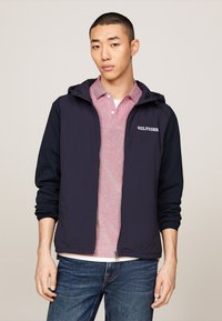 Giacca leggera navale con cappuccio e chiusura con zip, abbinata a una camicia a righe rosse e bianche con colletto. Il modello indossa jeans blu. Branding semplice visibile.