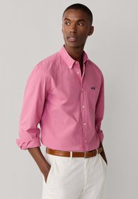 Mann trägt ein pinkfarbenes Hemd mit hochgekrempelten Ärmeln, weiße Hose und braunen Gürtel, schaut nach links, die Hände in den Taschen.