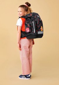 Un sac à dos noir avec des accents rouges et une bande réfléchissante, orné de patches. Un enfant portant un gilet orange et un pantalon rayé, se tenant de côté.