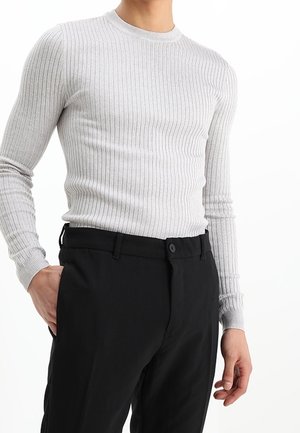 Mann trägt ein eng anliegendes, hellgraues, geripptes Langarmshirt, das in schwarze Anzughose gesteckt ist, Hand in der Tasche, vor weißem Hintergrund.