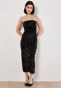 Schwarzes, trägerloses Pailletten-Kleid mit taillierter Silhouette, knielangem Saum und glatter Textur. Getragen mit spitzen schwarzen Absätzen.