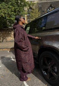 Naine, kes kannab mustrilist mantlit ja leopardimustriliste kingadega, avab tume Land Roveri tellishoone ette kaarakendega.