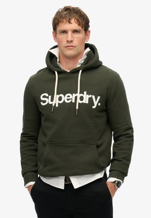 Man in een donkergroene "Superdry" hoodie over een witte blouse met kraag en zwarte broek, staande met handen in de zakken tegen een witte achtergrond.
