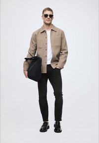 Világosbarna gombos dzseki egy fehér ing felett, fekete nadrág és fényes fekete cipő. Fekete táskát tart, sötét napszemüveget visel.