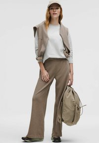 Beige sweatshirt med en ærmeløs, matchende vest, beige brede bukser, olivengrønne sneakers og en blød beige rygsæk.