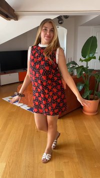 Vestido preto sem mangas com padrões de corações vermelhos, gola branca franzida e um corte solto. Modelo usando sandálias brancas, posando com as mãos levantadas.