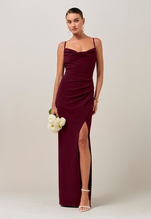 Kobieta w bezrękawnym bordowym sukni z rozcięciem na udzie trzyma białe kwiaty, ma na sobie białe szpilki i złotą biżuterię, stoi na beżowym tle.