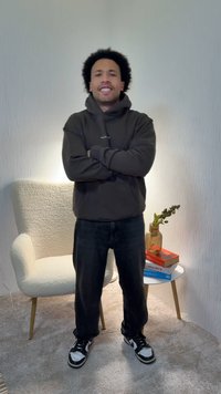 Mørk hettejakke, vide svarte jeans og svarte joggesko med hvite detaljer. Står i et godt opplyst rom med en beige stol og bøker.