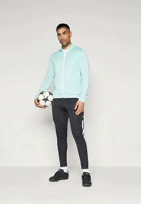 Light blue zip-up athletic jacket, black slim-fit pants with white stripes, and black sports shoes. Holding a black and white soccer ball.