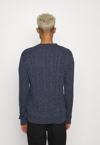 Pull en maille bleu foncé avec une texture côtelée, décolleté rond et manches longues. Le dos présente des détails de tricot en câble vertical.