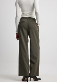 Olijfgroene wijde broek van stof, met een getailleerde pasvorm, riemlussen en achterzakken, gecombineerd met zwarte hoge hakken.