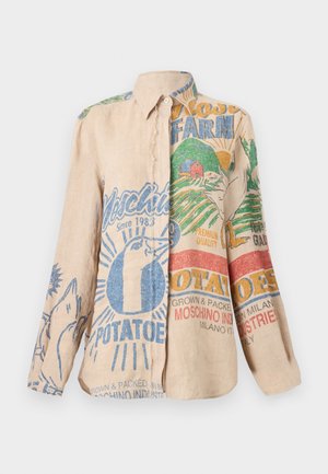 Nainen pukeutunut ylisuurikokoiseen beigenväriseen grafikalla varustettuun paitaan, jossa on värikäs maatila- ja perunaprintti, vaaleansiniset farkut ja mustat avoimet korkokengät, seisoo valkoista taustaa vasten.