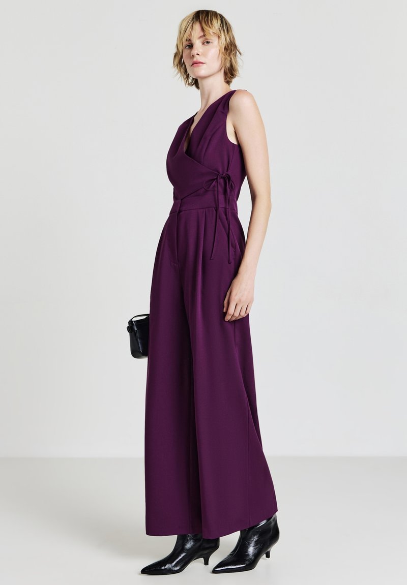 Frau in einem ärmellosen lila Jumpsuit mit weitem Bein, die eine schwarze Handtasche hält und schwarze Knöchelstiefel trägt, vor einem einfarbigen Hintergrund.