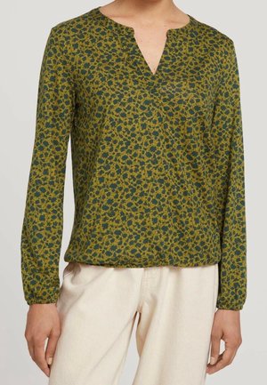 Blouse verte à manches longues avec un petit motif floral et un col rond fendu, portée avec un pantalon beige clair, montrée du cou jusqu'aux hanches.