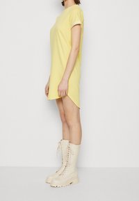 Robe jaune à manches courtes avec un col rond et une coupe droite, dotée d'un ourlet asymétrique. Associée à des bottes crème à lacets montantes jusqu'aux genoux.