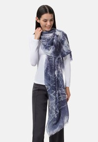 Sciarpa a motivo blu e bianco con bordi frangiati, realizzata in tessuto morbido, drappeggiata elegantemente attorno al collo del modello.