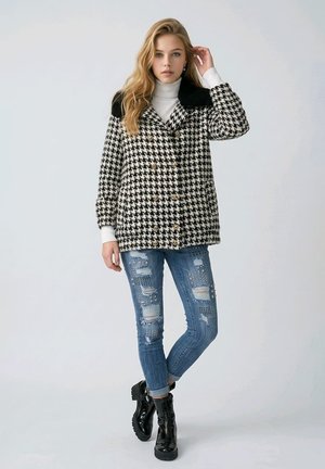 Mujer con abrigo de pata de gallo en blanco y negro, camiseta de cuello alto blanca, vaqueros azules desgastados con tachuelas y botines negros de charol.