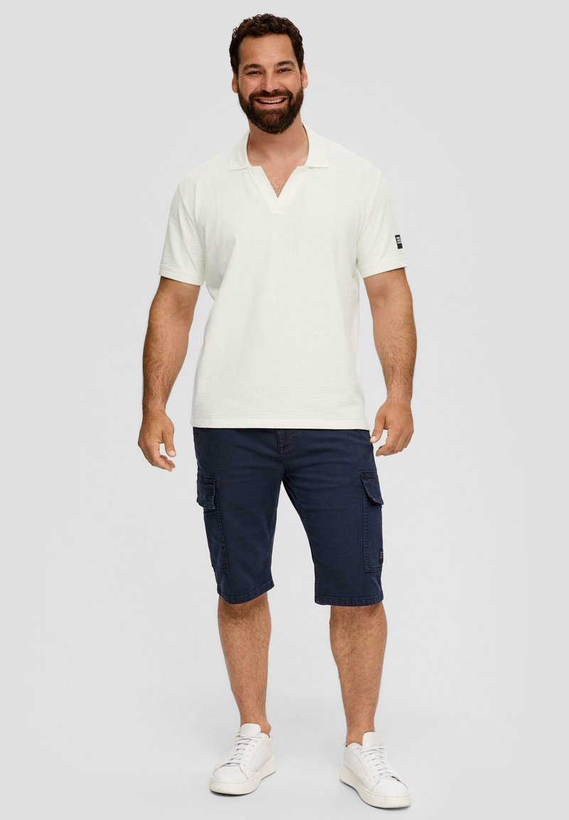 Weißes, strukturiertes Poloshirt mit V-Ausschnitt und kurzen Ärmeln, kombiniert mit marineblauen Cargo-Shorts und weißen Sneakers.