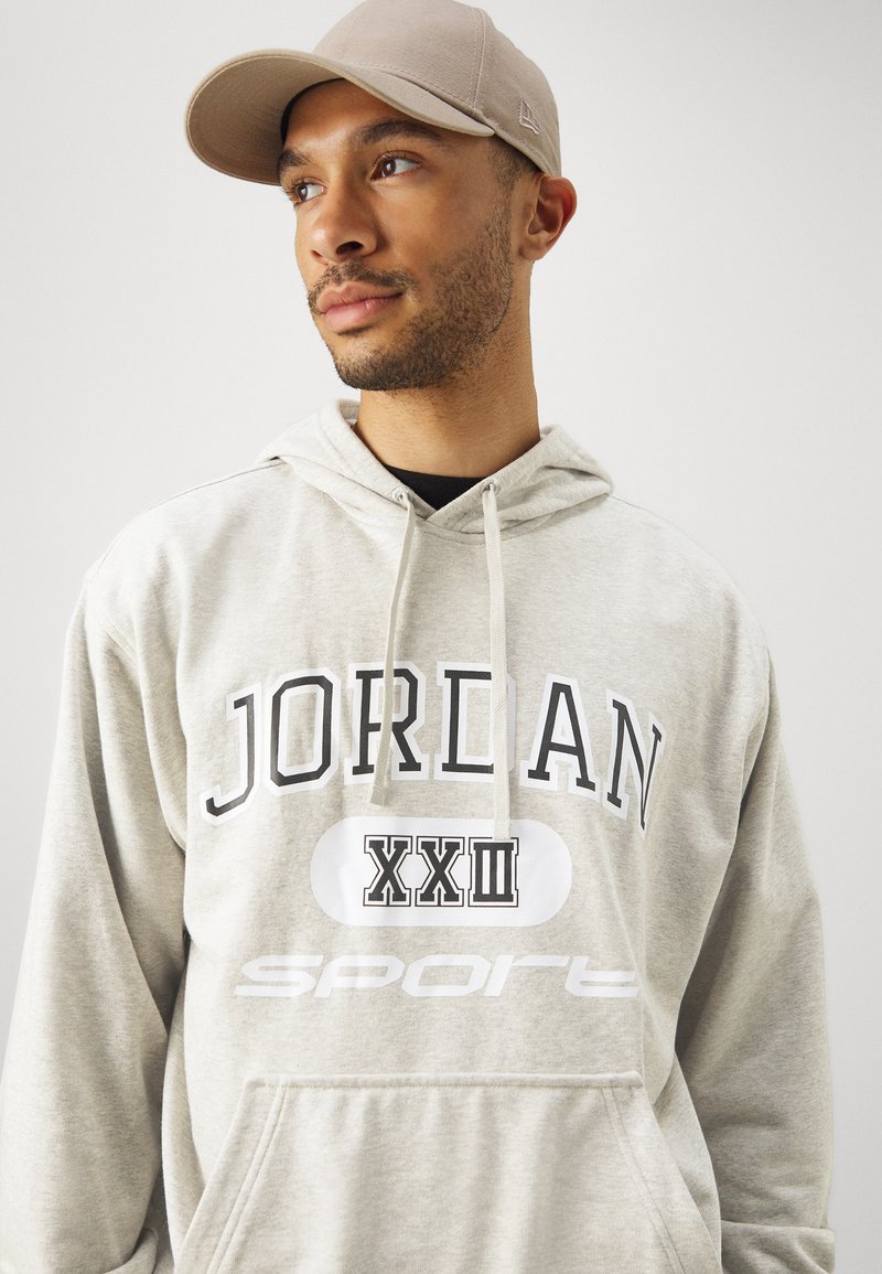 Hellgrauer Kapuzenpullover mit einer vorderen Kängurutasche, versehen mit schwarzem "JORDAN" und "XXIII" Schriftzug. Dazu wird eine beigefarbene Mütze getragen, die eine glatte Textur aufweist.