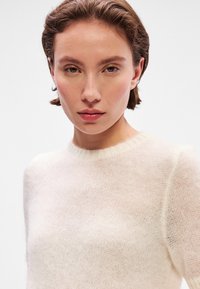 Ľahký, krémový sveter s okrúhlym výstrihom. Mäkka, textúrovaná tkanina s jemne priesvitným vzhľadom a rebrovanými detailmi na golieri.