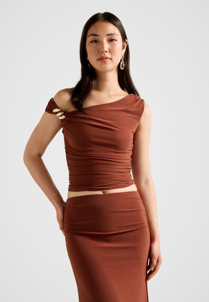 Γυναίκα που φοράει τοπ ώμου σε σκουριά απόχρωση με ρυτιδώσεις και χρυσά τσιμπιδάκια σε ένα μανίκι, καθώς και εφαρμοστή φούστα στο ίδιο χρώμα.