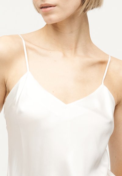 Femme portant un haut caraco en satin blanc avec de fines bretelles spaghetti, ligne de la mâchoire et clavicules visibles sur un fond uni.