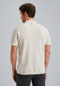 Helles beiges Poloshirt mit kurzen Ärmeln aus strukturiertem Strick, das ein kleines rundes Logo auf der Rückseite nahe dem Kragen trägt.