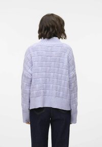 Lila stickad tröja med en texturerad korg vävdesign, hög krage och ribbad nederkant, bärs med mörka jeans, sedd bakifrån.
