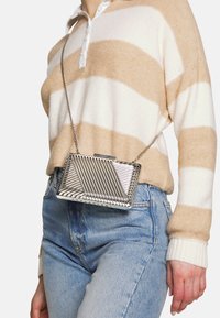 Metaliczna srebrna torebka kopertówka o geometrycznym wzorze, z ukośnymi liniami i łańcuszkowym paskiem, noszona na tle paskowanego swetra w odcieniach beżu i bieli.