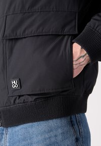 Hånd i lommen på sort HUGO-jakke, der viser ærmekanten, nederste lomme med logo og blå denimjeans. Synlig tatovering på hånden.