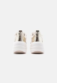 Ζευγάρι μπεζ αθλητικά παπούτσια με μεταλλικές χρυσές λεπτομέρειες στη φτέρνα και χοντρές λευκές σόλες, απεικονιζόμενα από πίσω σε απλό ανοιχτό φόντο.