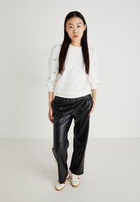 Sudadera blanca con detalle de ojales en las mangas, combinada con pantalones de cuero sintético de pierna ancha negros y zapatillas blancas con acentos rojos.