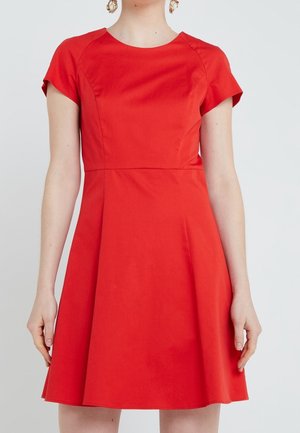Mujer con un vestido rojo de manga corta, largo hasta la rodilla, con cuerpo ajustado y falda acampanada sobre un fondo liso.