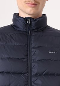 Marine pufferjack met een hoge kraag, doorgestikte textuur, ritssluiting aan de voorkant en subtiele merkbranding. Het heeft een gladde, glanzende buitenkant.