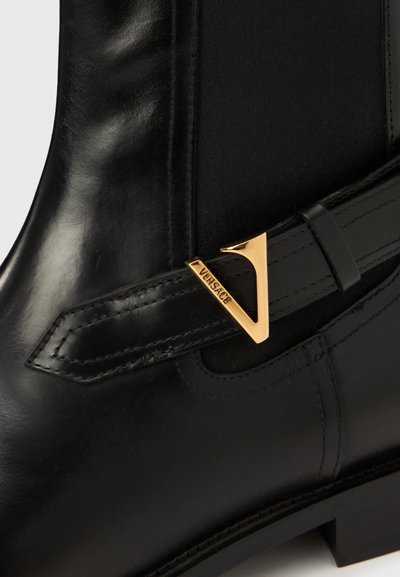 Černá kožená kotníková bota s dekorativní zlatou sponou a kontrastním elastickým panelem. Design je elegantní, s hladkou texturou a pevnou podešví.