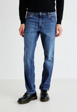Modri jeans iz denima s ravnim krojem, ki vključujejo zanke za pas, sprednje žepe in subtilen vzorec pranja. Oblečeni z črnimi škornji.