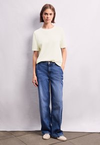 Lichtgele, kortärmelig gebreide top gecombineerd met blauwe jeans met brede pijpen en beige schoenen. Gladde textuur met eenvoudige designelementen.