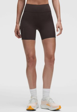 Donna che indossa pantaloncini sportivi neri a vita alta, calzini bianchi alla caviglia e scarpe da corsa bianche e arancioni su uno sfondo uniforme.