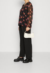 Svart blommig blus med genomskinliga långa ärmar, matchad med svarta vida byxor och svarta platåskor. Håller en liten off-white handväska.