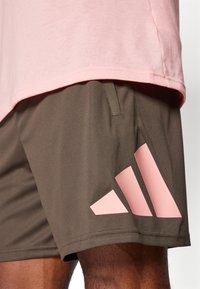 Pantalones cortos deportivos marrones con una textura suave, que presentan un destacado logo de tres rayas rosas en el lado izquierdo y un bolsillo lateral.