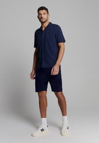 Marineblauwe overhemels met korte mouwen en een platte kraag, gecombineerd met bijpassende marineblauwe shorts. Model draagt witte sneakers met marineblauwe accenten.