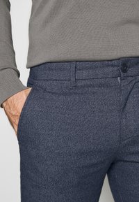 Mörkblå texturerade byxor med en slät finish, med en framficka och knappstängning. Grå tröja delvis synlig ovanför.