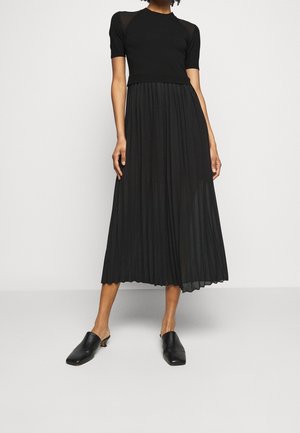 Woman wearing a black short-sleeve top with a sheer pleated midi skirt and black heeled mules, standing against a plain light background.
