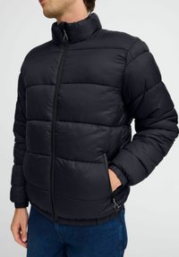 Svart polstret jakke med høy krage, frontglidelås og sidelommer. Har en quiltet design og elastiske mansjetter. Komplementerer denimbukser.